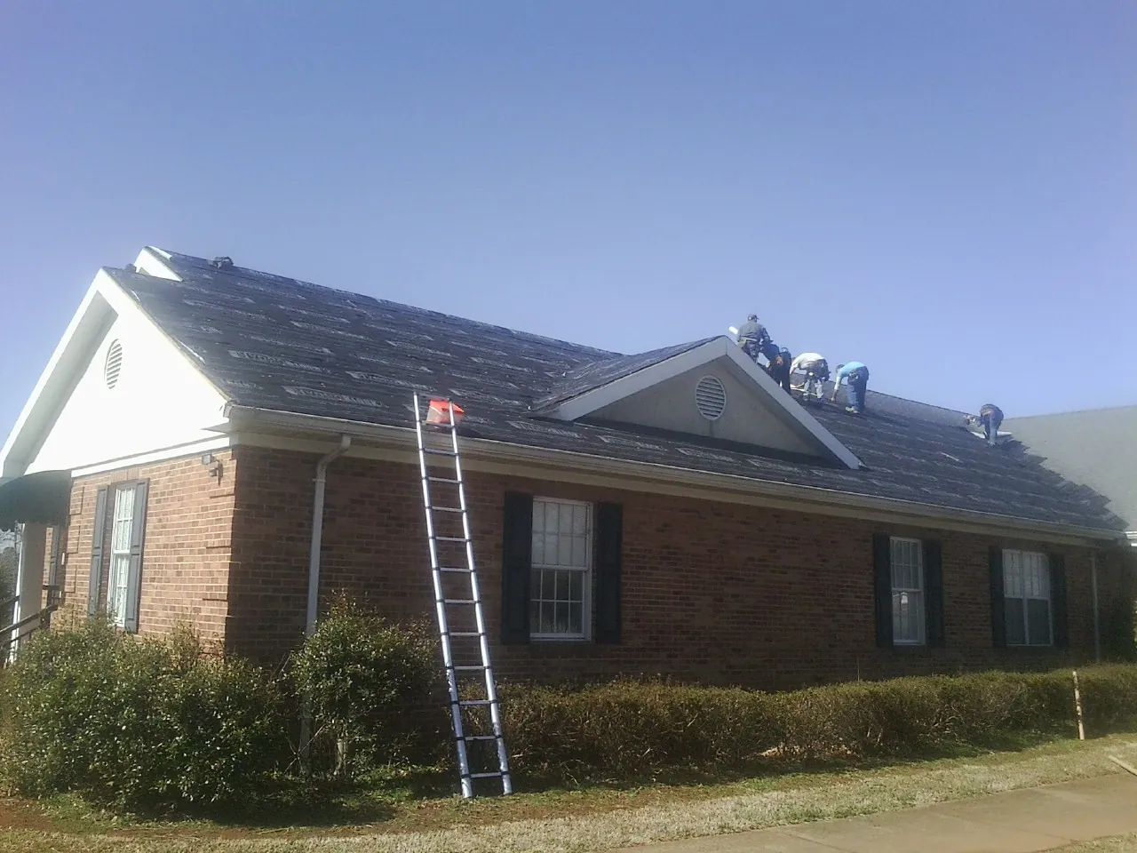 Crew tearing shingles off a brick ranch roof with a ladder set — Emergency Roof Repair work by our crew in La Fontaine