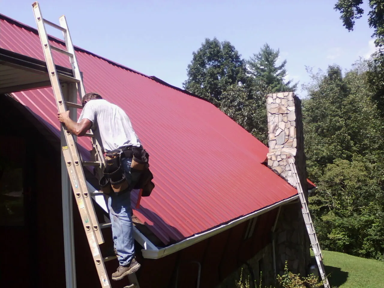 Roofer on a ladder beside a red metal roof and stone chimney — Soffit & Fascia Repair by Roofing Brothers in La Fontaine